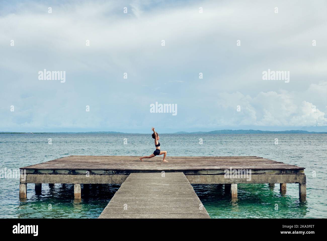 Distant female in black activewear performing Ashta Chandrasana with arms up against peaceful sea Stock Photo