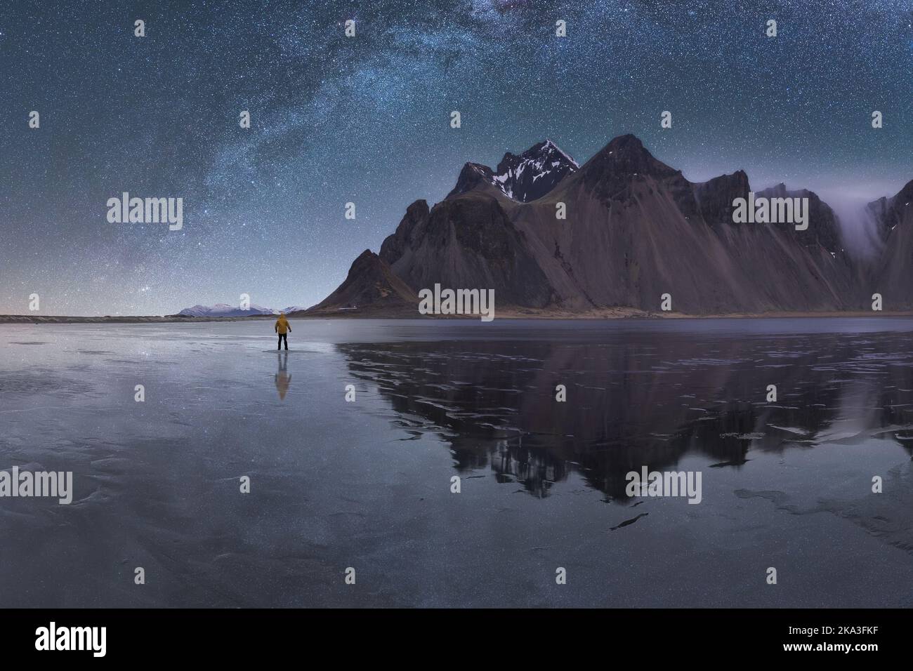 Anonymous tourist standing on reflection surface on wet shore near calm sea and rough mountains