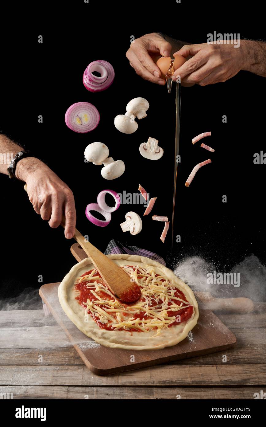 Crop hands breaking egg and spreading sauce and cheese with spatula pizza while mushrooms and onion with bacon falling on top against black background Stock Photo