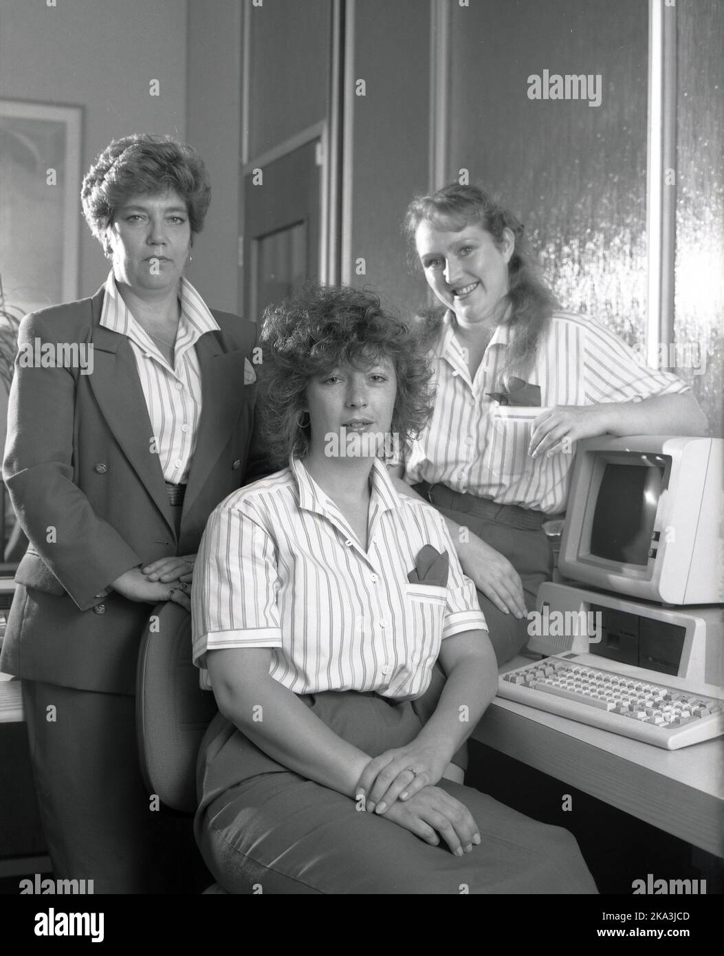 1989, historical, inside an office, three female employees, wearing company uniform clothing, gathered for a photo by a computer of the era, a small desktop IBM personal computer, consisting of display monitor, computer and keyboard, England, UK. Office manager in a jacket Stock Photo