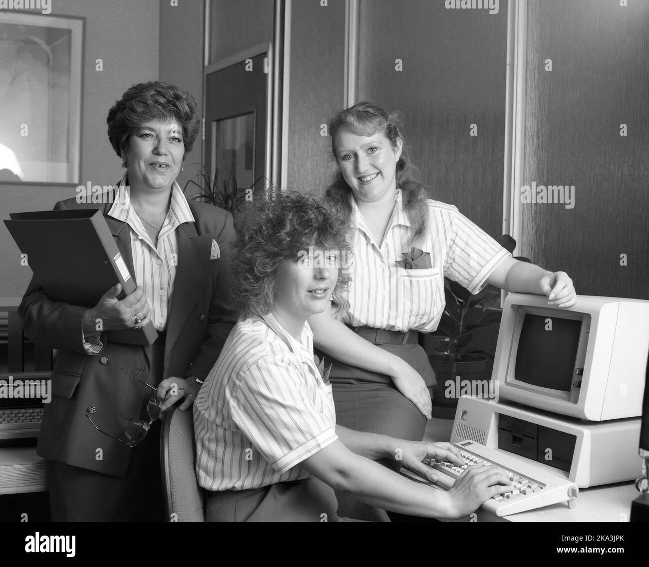 1989, historical, three office female staff by a computer of the era, England, UK. Manager in jacket behind, with folder, labeled 'House Rules'. One of the women is sitting at a keyboard infront of an IBM personal computer of the era, consisting of a separate display monitor and computer. Stock Photo