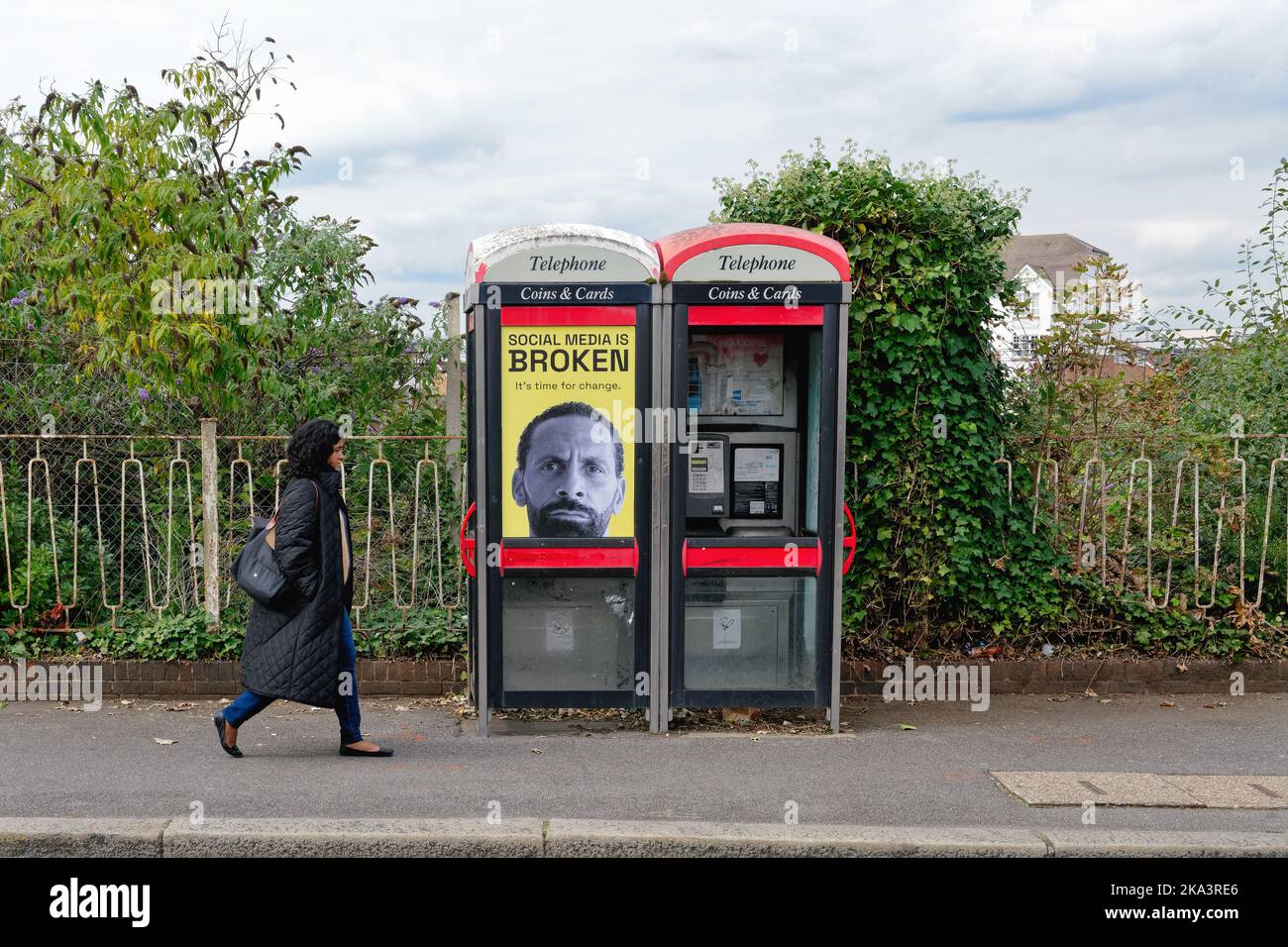 A young Asian woman walking by two derelict BTphone boxes that has an poster featuring Rio Ferdinand stating 'Social Media is Broken' Hounslow London Stock Photo