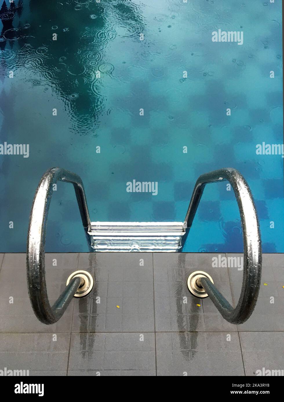 A vertical high angle shot of a metal staircase inside a blue pool with raindrops falling on it Stock Photo