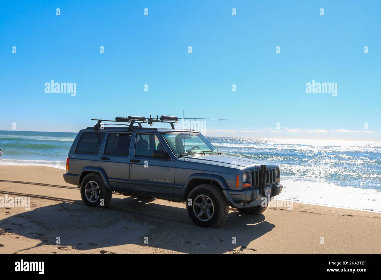 https://c8.alamy.com/comp/2KA3TBF/the-jeep-cherokee-driving-on-a-sunny-beach-next-to-the-ocean-with-fishing-rods-on-the-roof-montauk-2KA3TBF.jpg