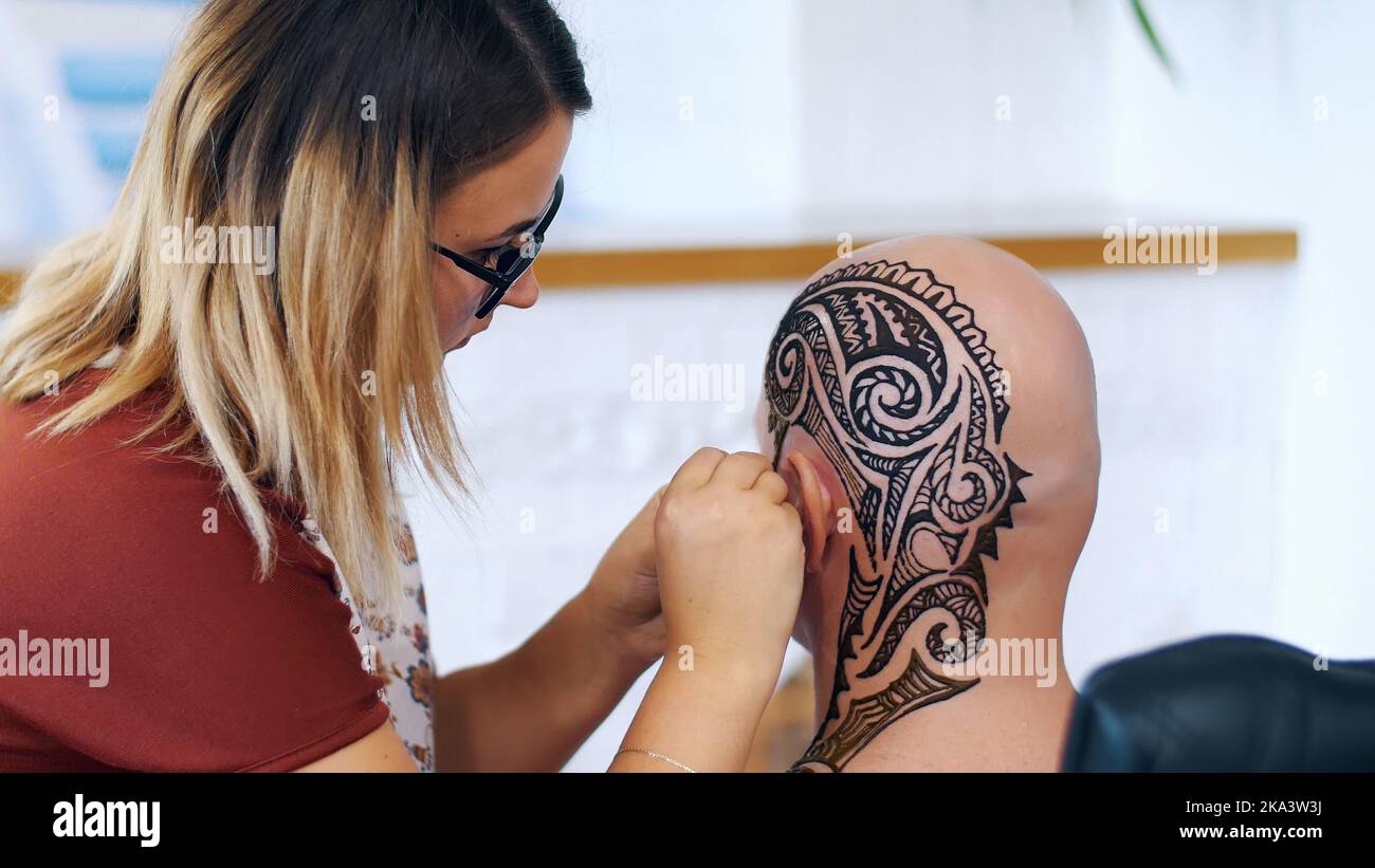 A girl, tattoo master, mehendi artist makes drawing of henna tattoo on scalp of bald Caucasian man, shoulder, neck.The process of applying henna from a tube to the skin. High quality photo Stock Photo