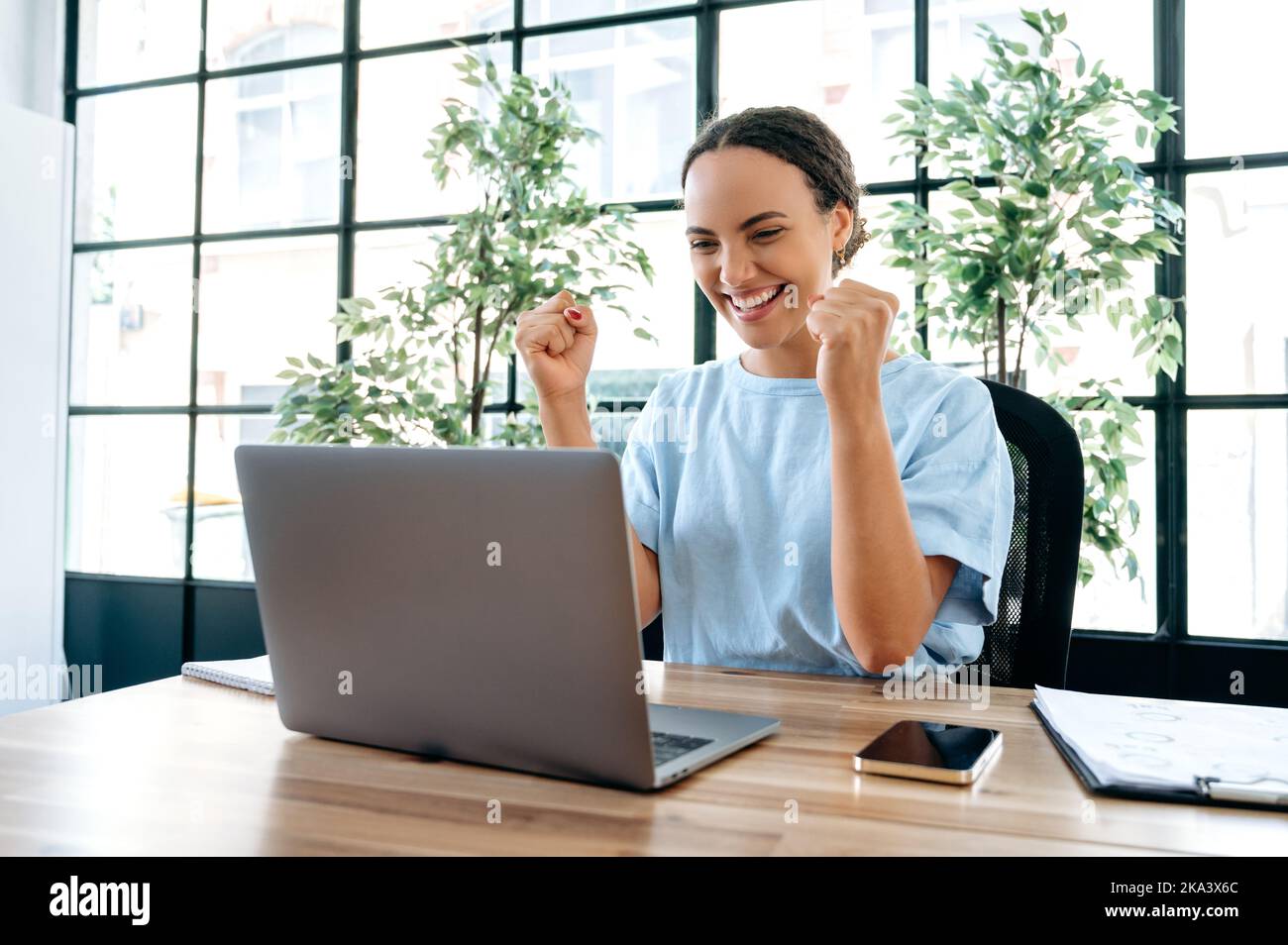 Amazed happy brazilian or hispanic business woman, sit at a desk with laptop in modern office, rejoicing in success, big profit, celebrate deal, job promotion, gesturing fists, looks at camera, smiles Stock Photo