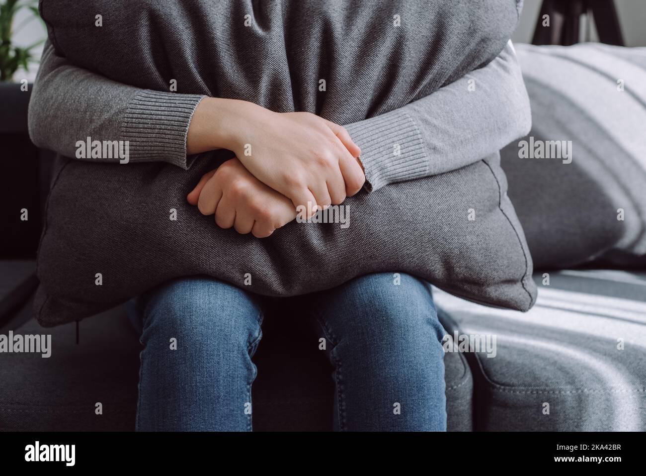 Unhappy lonely depressed young woman at home sitting on couch hiding her face big grey pillow, crying feeling stressed, mourning, suffering from negat Stock Photo