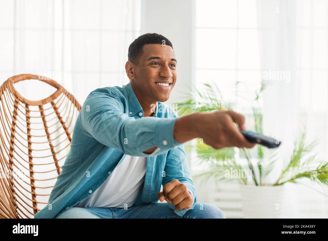Smiling millennial african american guy with remote control switches tv, enjoy spare time, sits on chair Stock Photo