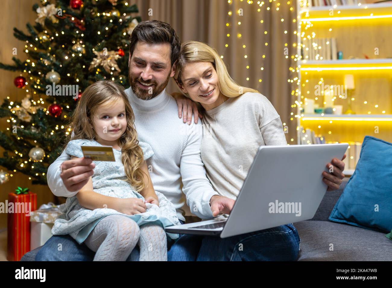 Happy Christmas family doing online shopping sitting at home on sofa, couple husband wife and daughter with laptop and bank credit card having fun choosing Christmas gifts near Christmas tree. Stock Photo