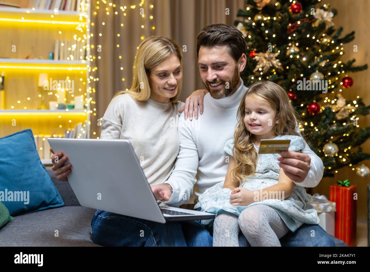 Happy Christmas family doing online shopping sitting at home on sofa, couple husband wife and daughter with laptop and bank credit card having fun choosing Christmas gifts near Christmas tree. Stock Photo