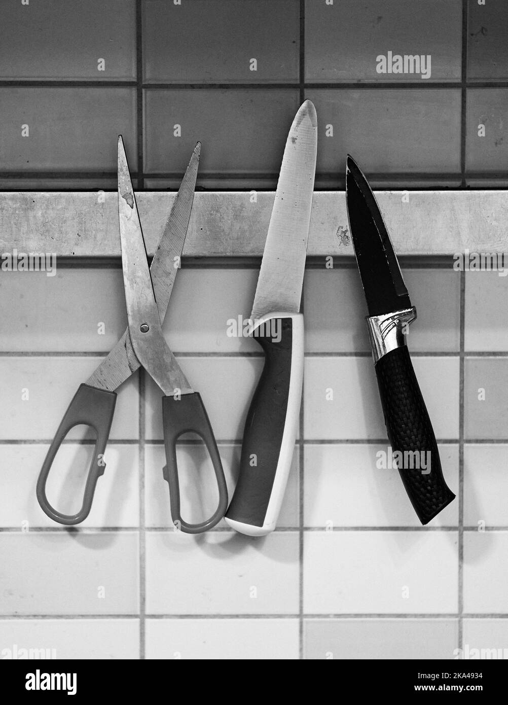 A grayscale shot of scissors and two kitchen knives hanging on the wall Stock Photo