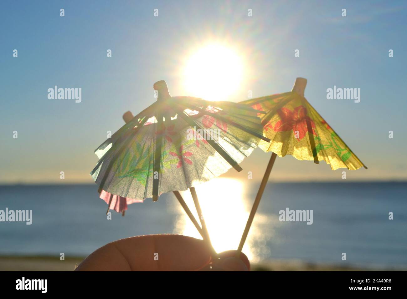 Man holding in hand three paper umbrellas cocktail decorations on background of blue sky, brightly shining sun and sunny path on surface of blue sea. Concept of summer, sea, vacation, travel, tourism Stock Photo
