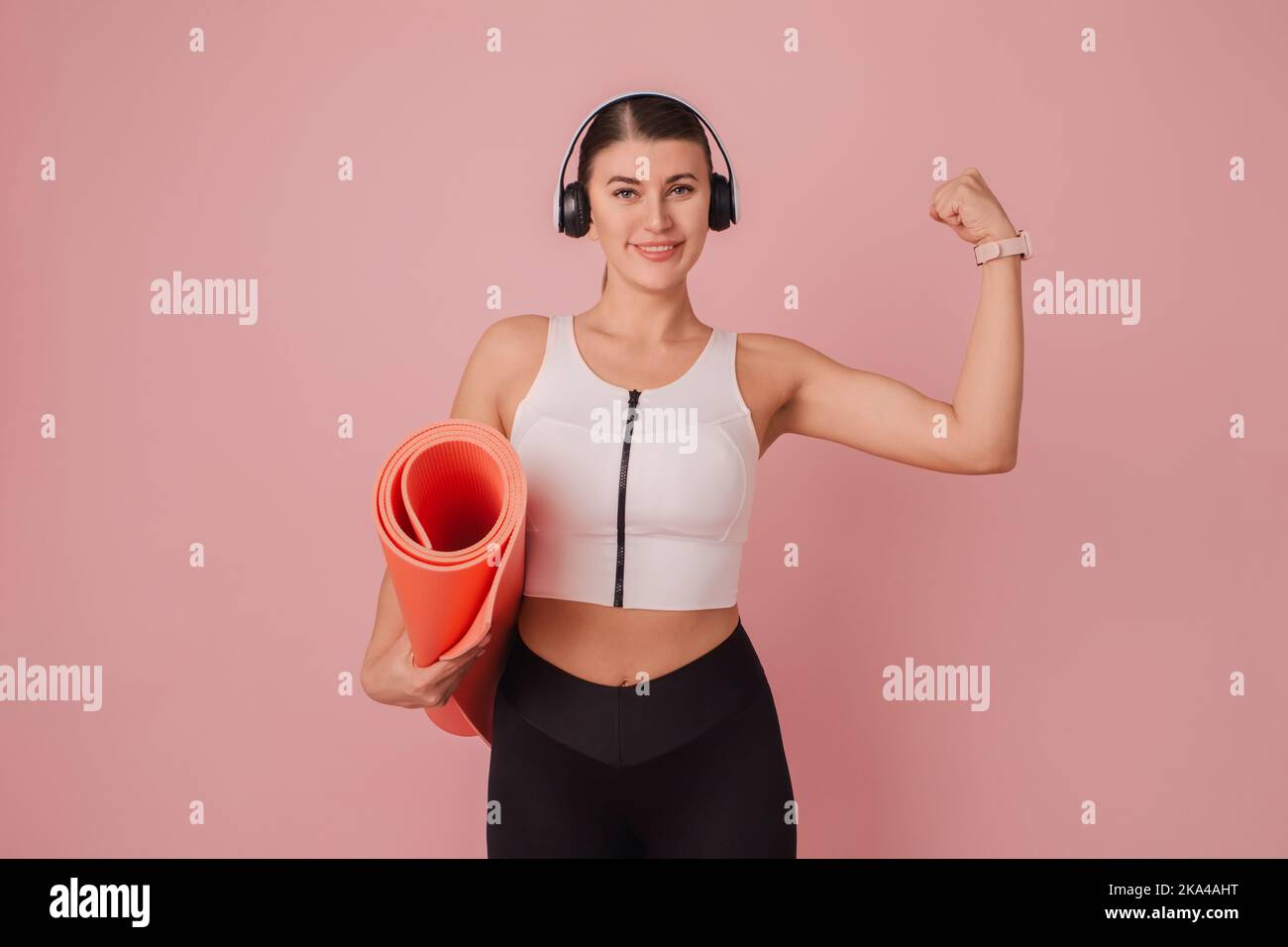 Sport motivation concept. Young smiling woman standing in headphones with fitness mat on pink background Stock Photo