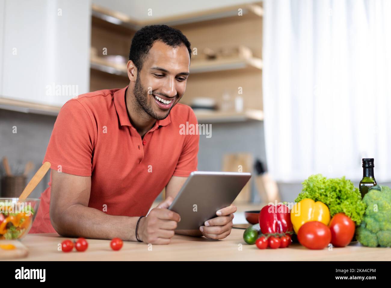 Smiling young black guy in red t-shirt watch video on tablet at table with organic vegetables Stock Photo