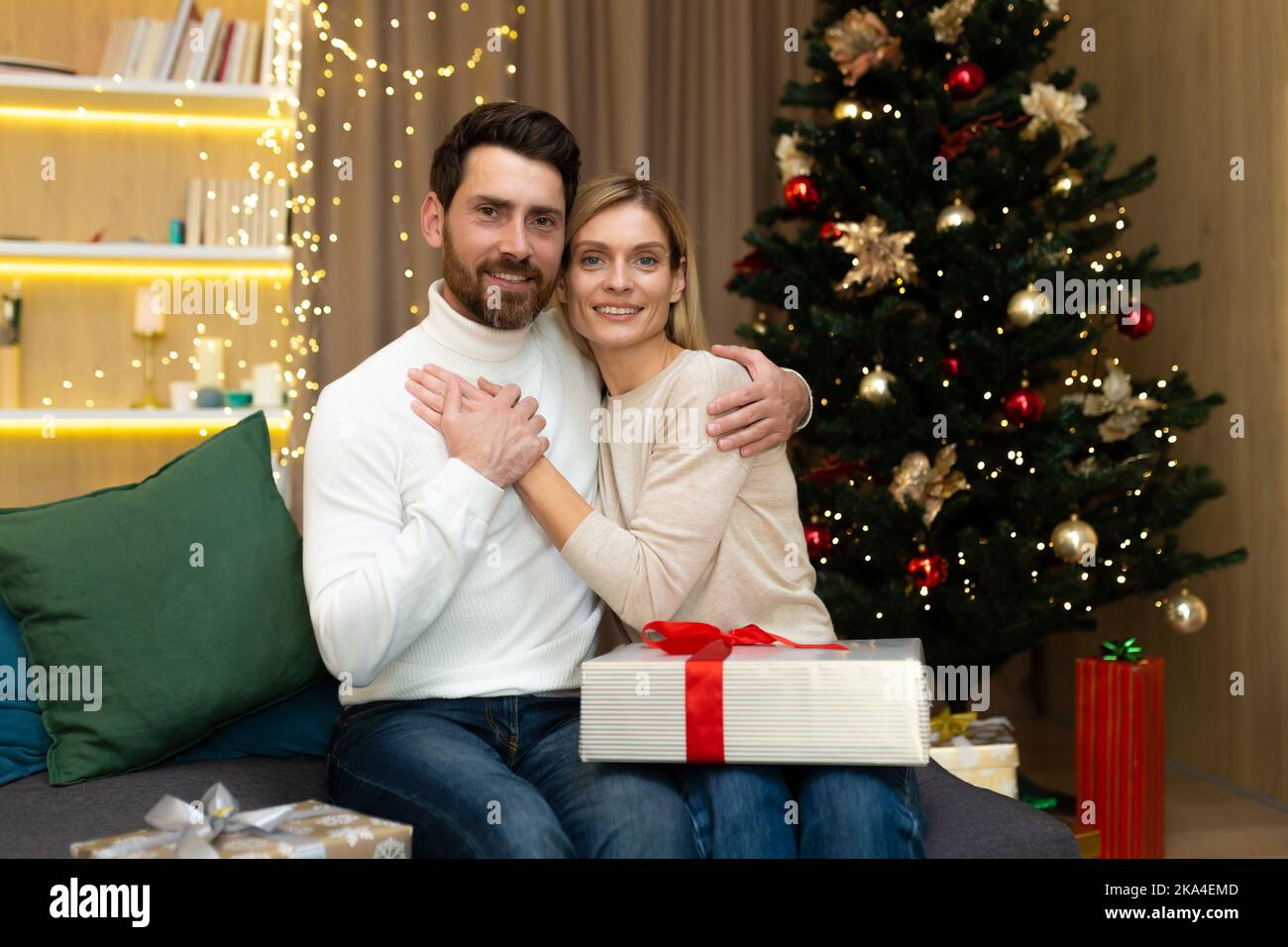 Portrait of happy married couple on Christmas, husband and wife sitting with New Year's gifts at home on sofa and looking at camera, smiling near Christmas tree. Stock Photo