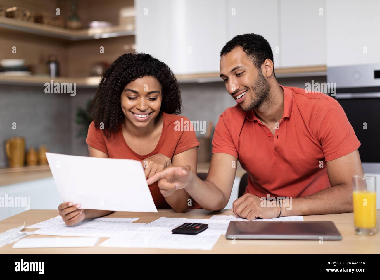 Cheerful young black female in red t-shirt shows documents to man with pc, check accounts use banking Stock Photo