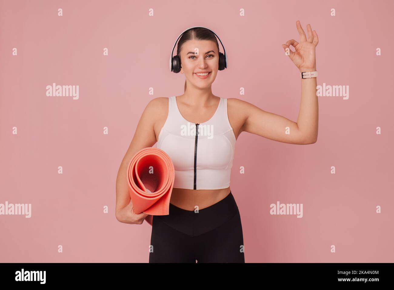 Happy fitness girl. Smiling woman standing in headphones with fitness mat on pink background with OK gesture Stock Photo