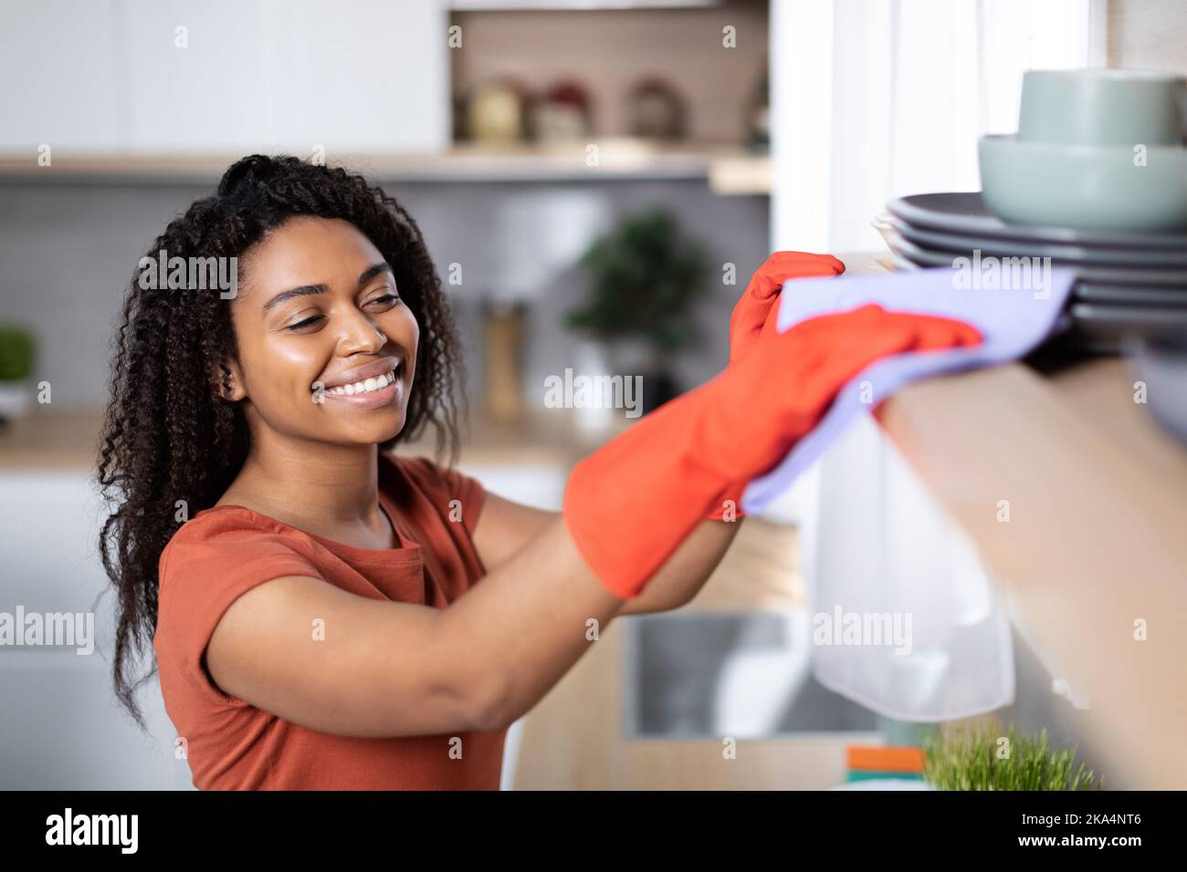 Cheerful pretty young black woman in red t-shirt and rubber gloves dusts off from wooden shelf Stock Photo