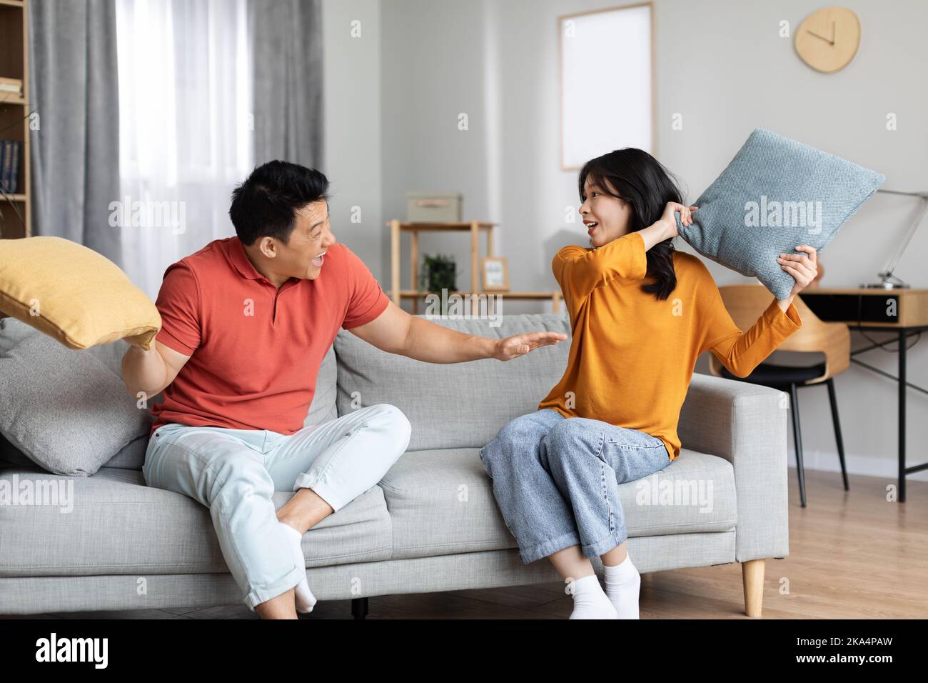 Asian spouses in homewear sitting on couch, having pillow fight Stock Photo