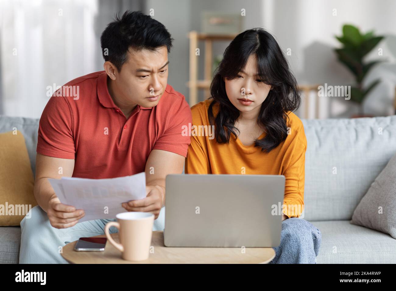 Upset asian couple checking bills, using laptop, home interior Stock Photo