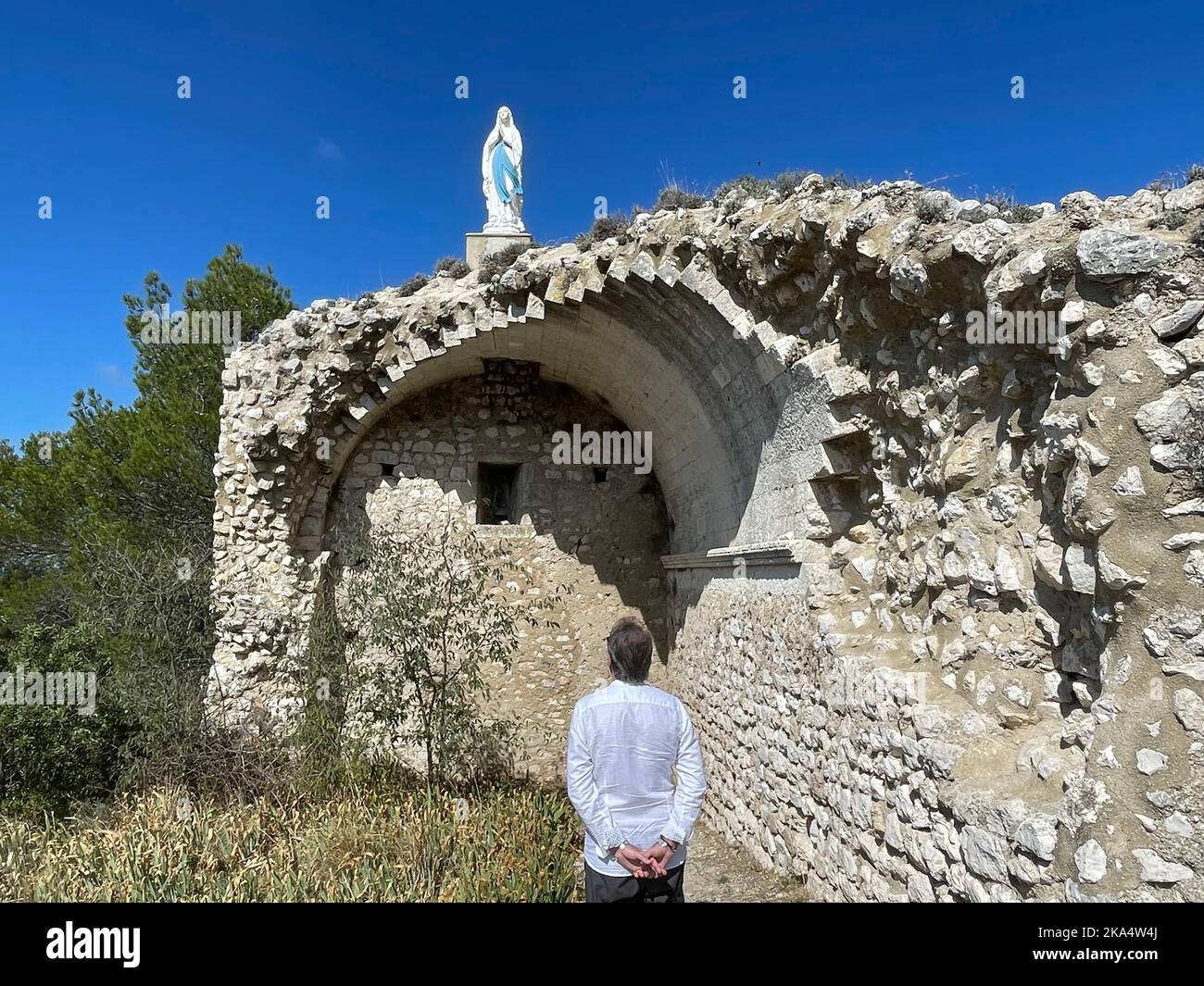 Rear view of a man with his hands clasped behind his back looking up at La statue de la vierge juchee, Eygalieres, Bouches-du-Rhone, Provence-Alpes-Cote d'Azur, France Stock Photo