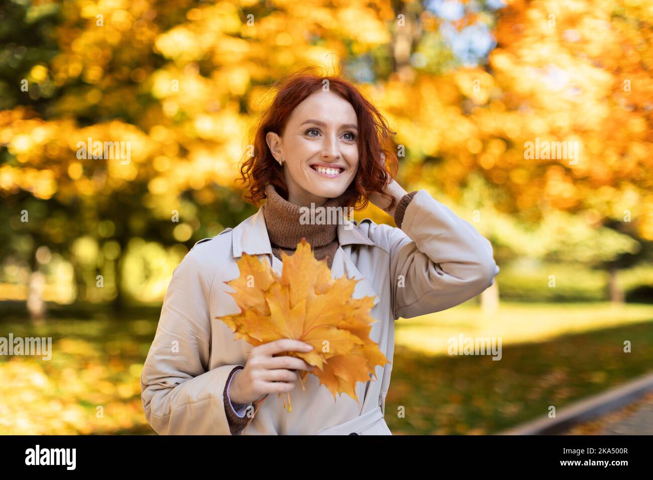 Urban lifestyle. Glad pretty smiling young european red-haired woman in raincoat hold yellow leaves Stock Photo
