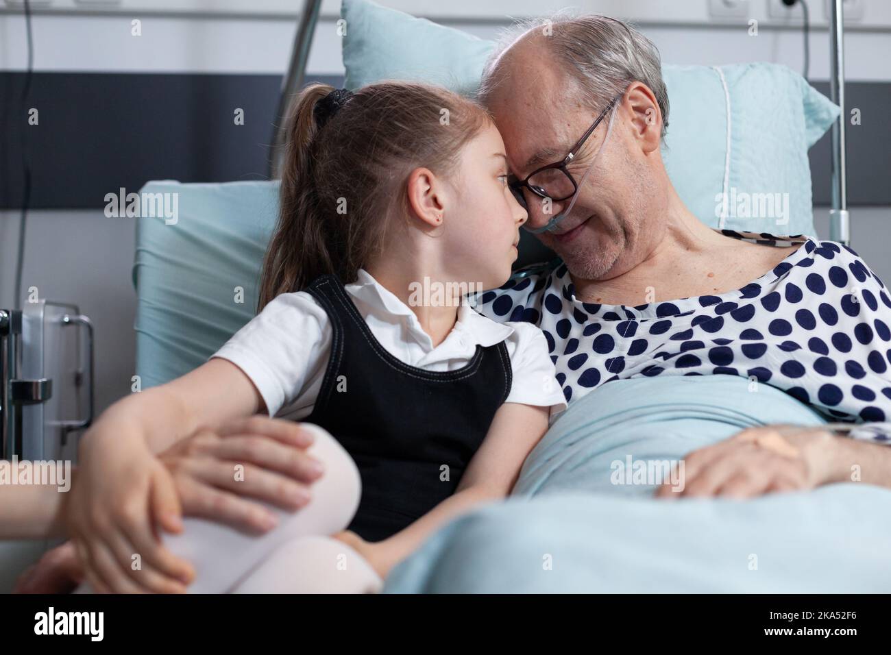 Elderly recovering patient happily cuddling infant granddaughter in hospital observation room bed. Senior man enjoying company of relatives visit in geriatric clinic room. Stock Photo