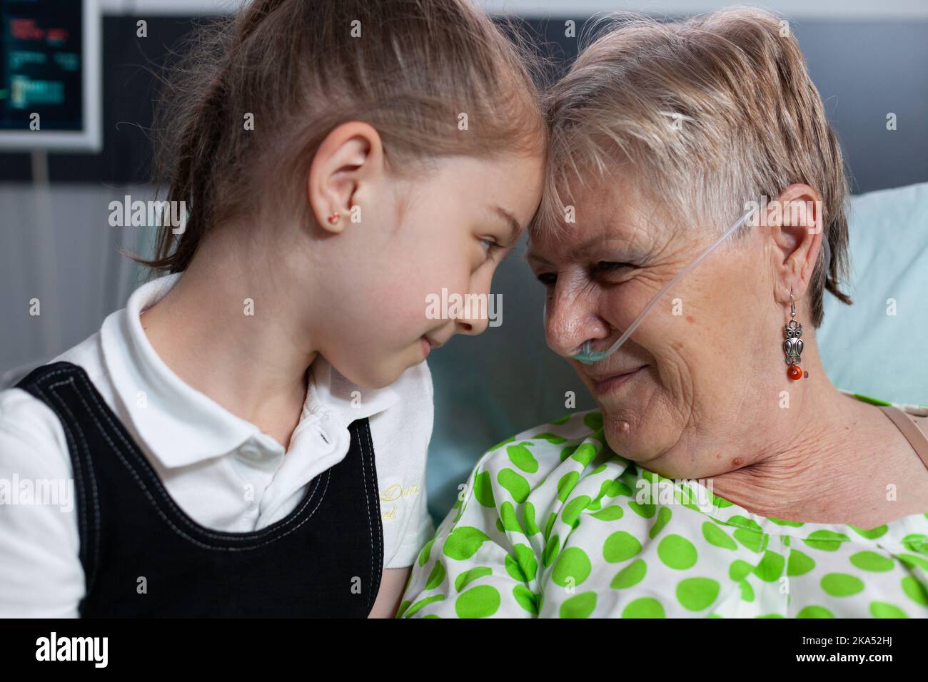 Elderly woman lying in hospital bed with breathing aids showing affection to little girl. Grandmother and granddaughter bumping heads in love at medical clinic for elderly people recovery room. Stock Photo