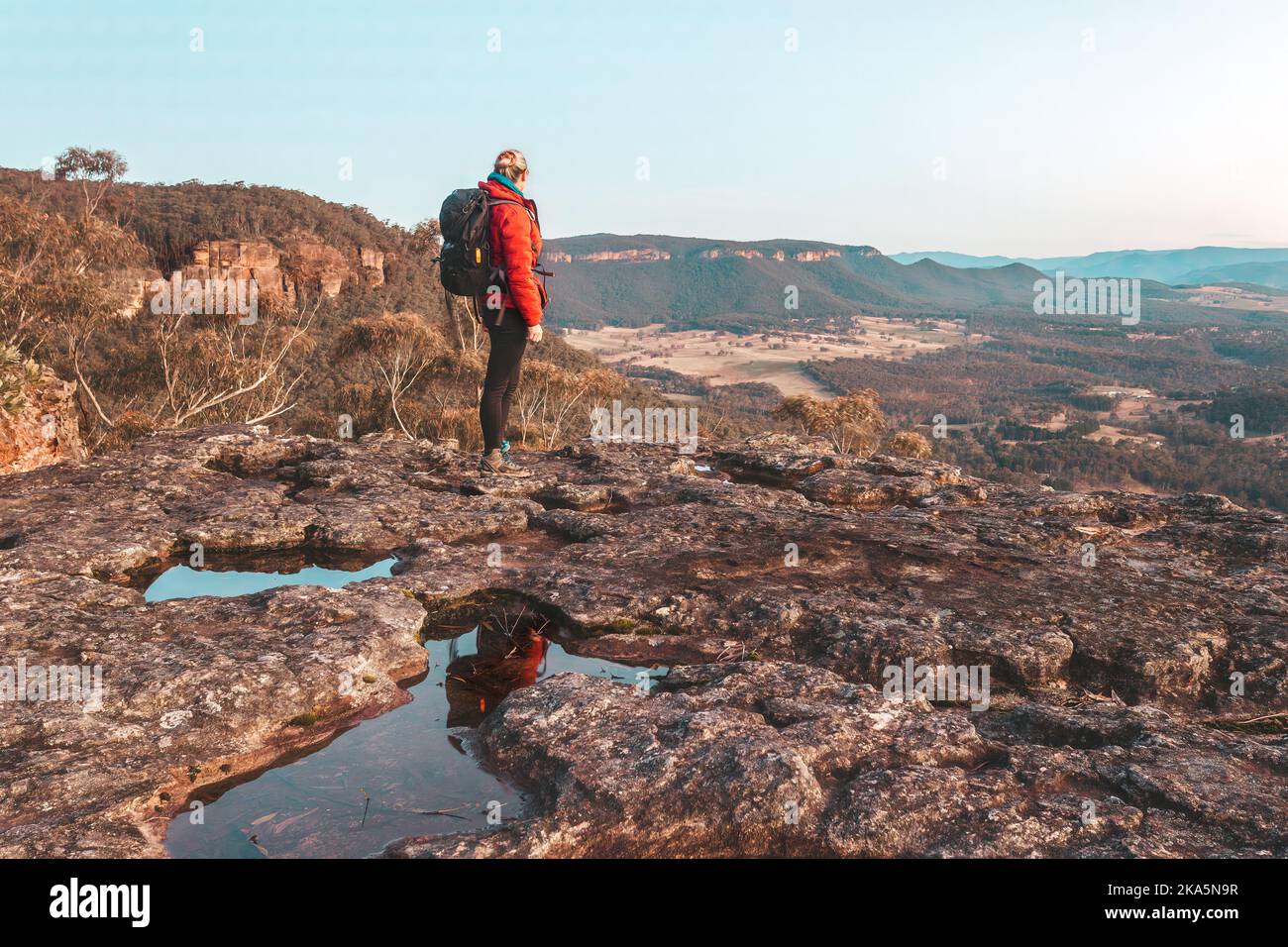 Female hiker at to the top of a rugged cliff face overlooking a valley and other mountains in the distance Stock Photo