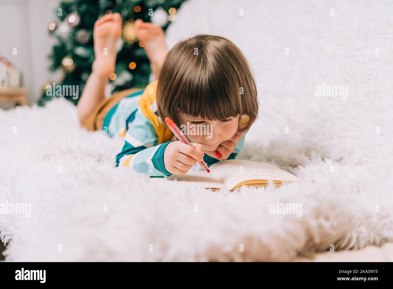 Smiling Kid boy lying on couch and writing the letter to dear Santa at home near Christmas tree. Child wish list. Dreams of a Christmas gifts. Merry Stock Photo