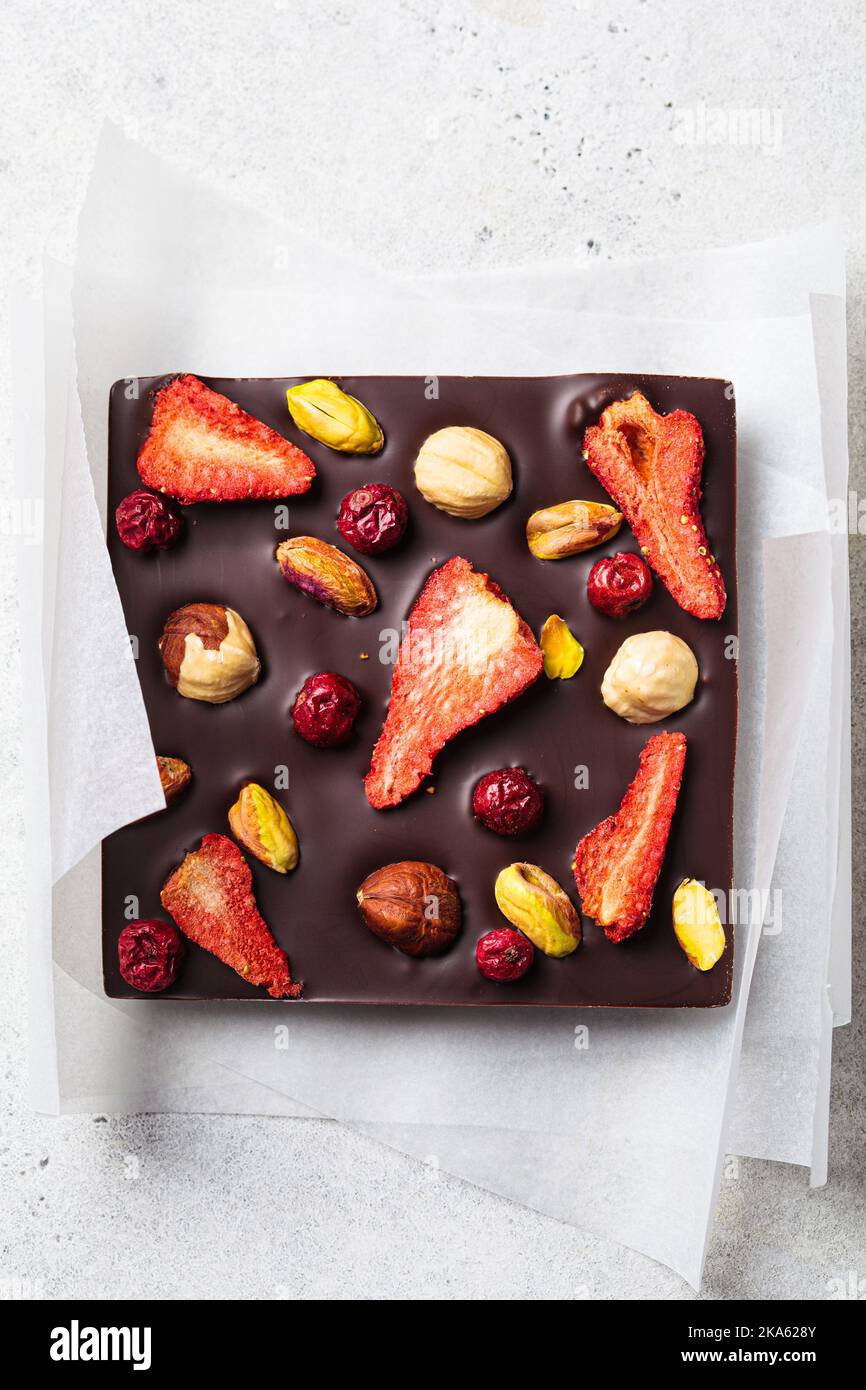 Bar of homemade dark chocolate with berries and nuts on white paper, top view. Stock Photo