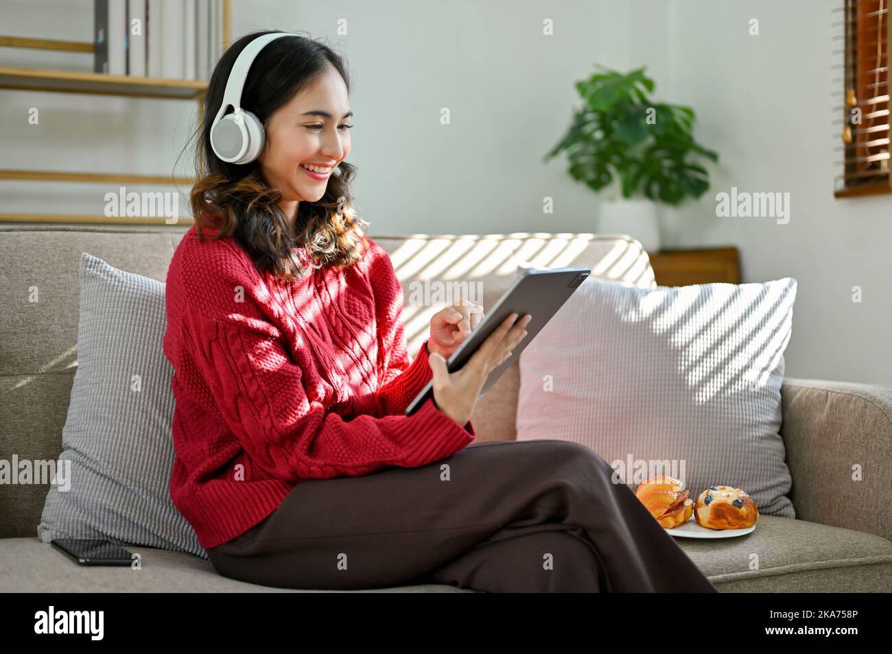 Cheerful and beautiful young Asian female in red sweater, wearing headphones, using her digital tablet, sitting on sofa in living room. Stock Photo