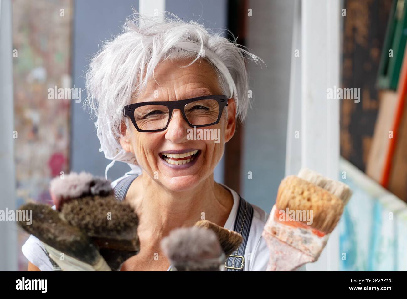 smiling happy older woman portrait, proud artist, in her fifties with grey hair and black glasses and many paintbrushes, copy space Stock Photo