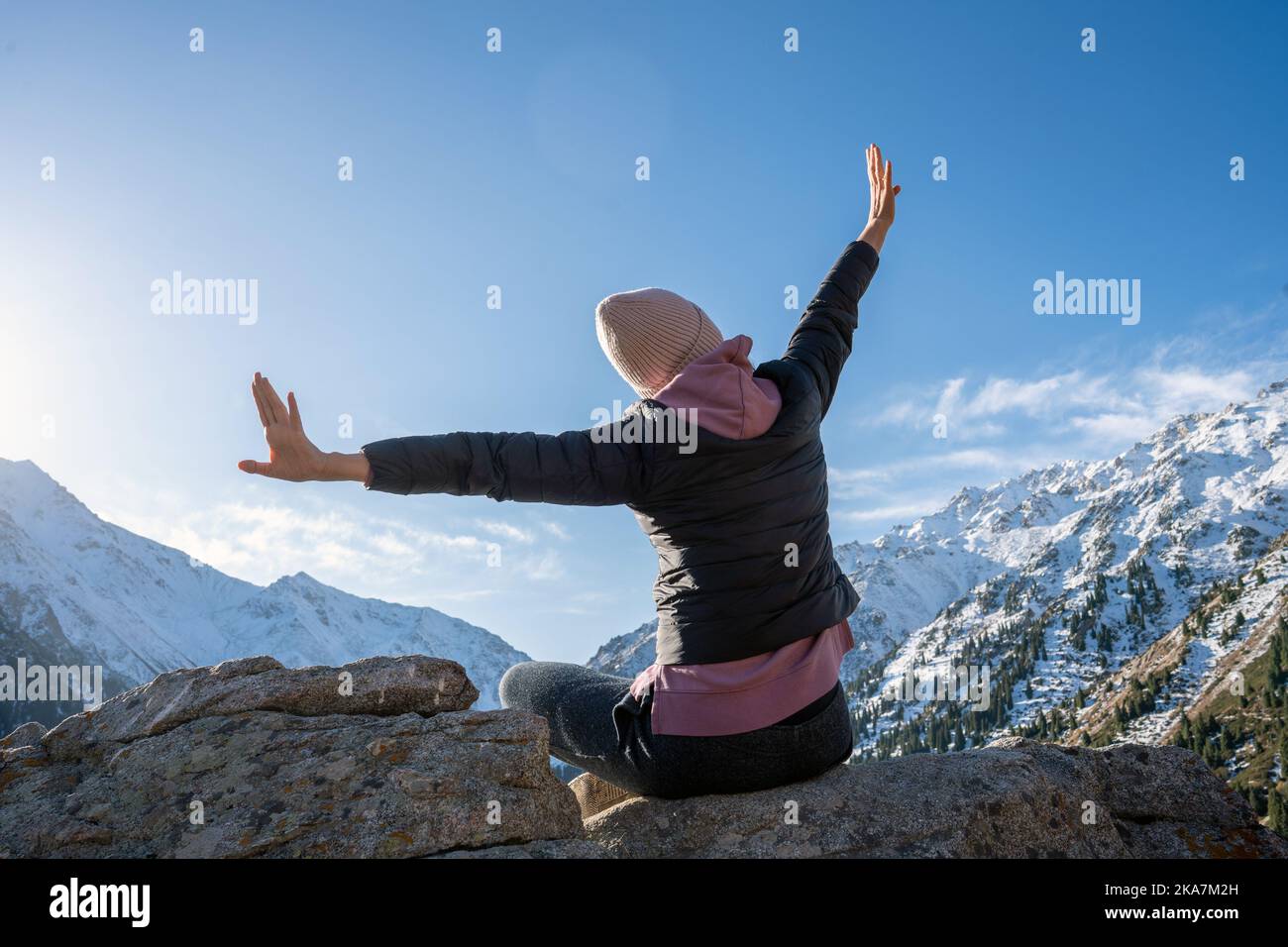 Elated woman with hands outstretched like wings on a vantage in snowy mountains on a sunny day. She is sitting on a boulder. Stock Photo