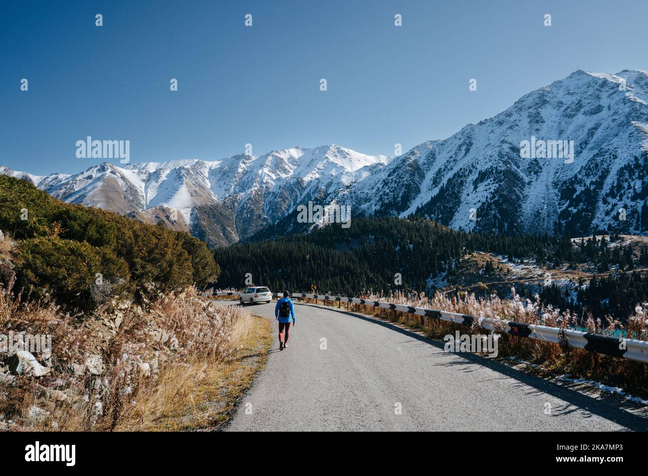 Longshot of a man walking on the road surrounded by beautiful mountain scenery. Trees by his sides and snowy peaks in the distance. Stock Photo