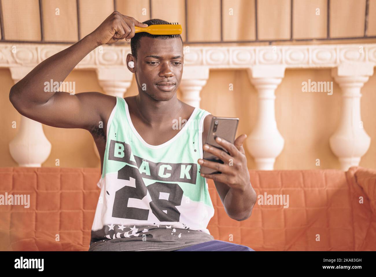 Black man in sports tank top combing hair and looking at screen of smartphone while sitting on bench in cafeteria Stock Photo