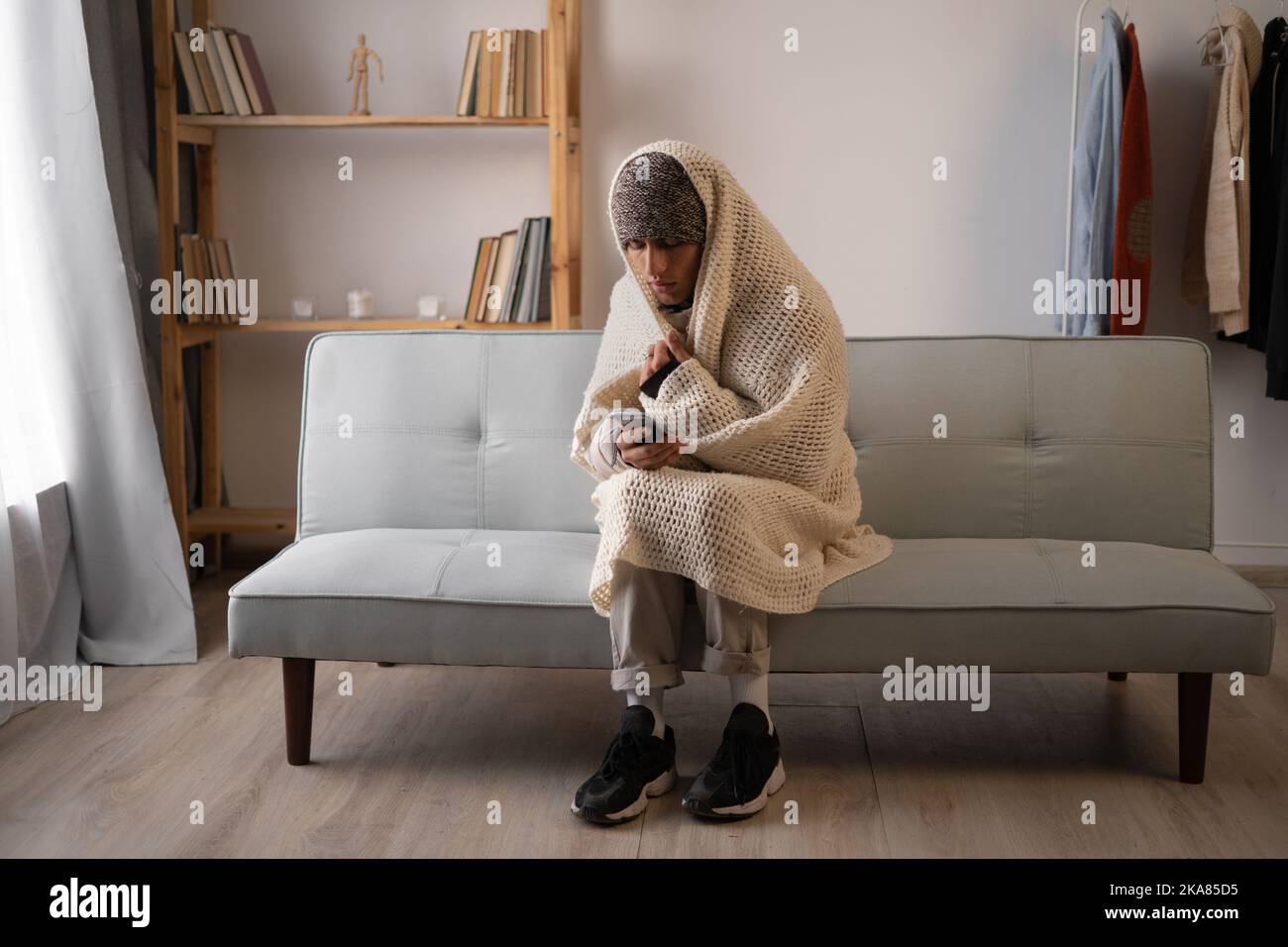 Unwell man renter in blanket sit in cold living room use mobile phone.suffer from lack of heat and power. Unhealthy young man struggle from chill Stock Photo