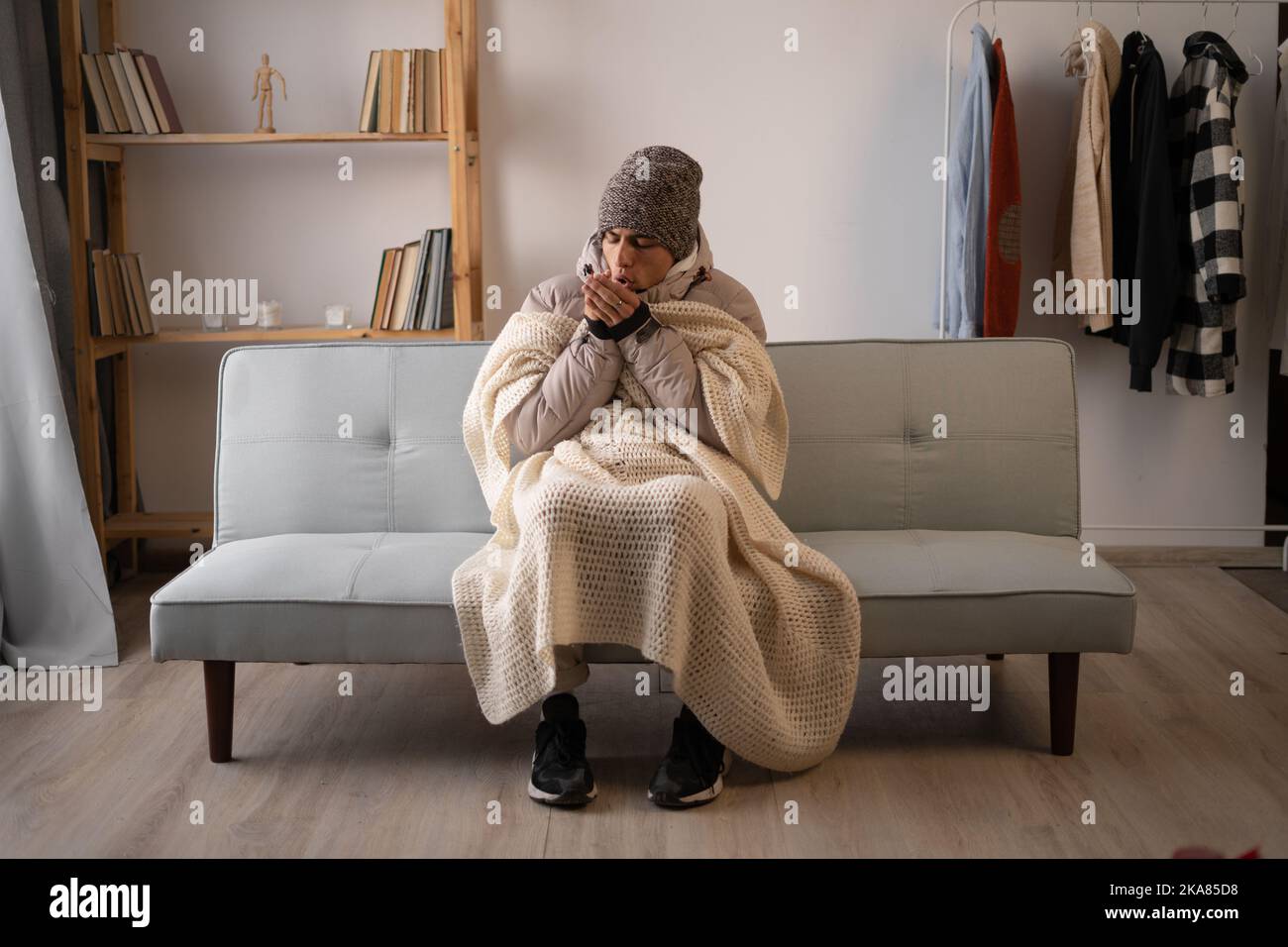 Unwell man in hat and blanket sit in cold living room suffer from heat