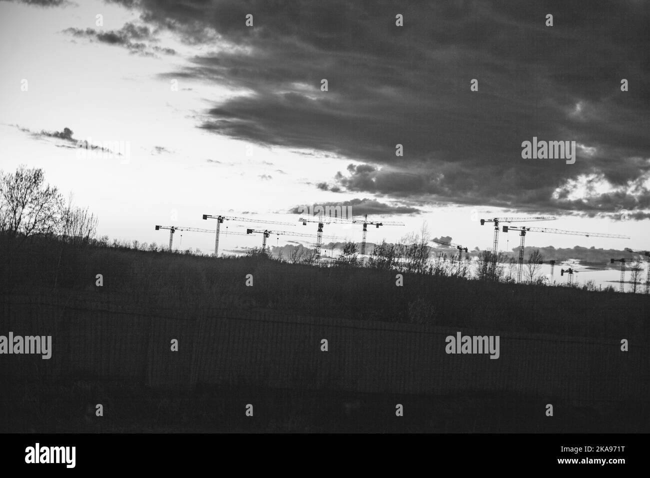 A grayscale of construction cranes in the countryside under the cloudy ...