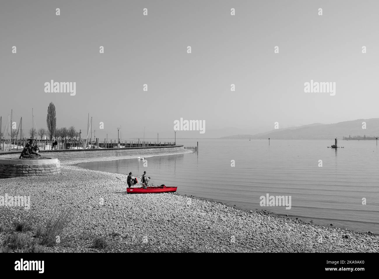 A grayscale shot of the rocky beach of the sea with a red boat and passengers on the shore Stock Photo