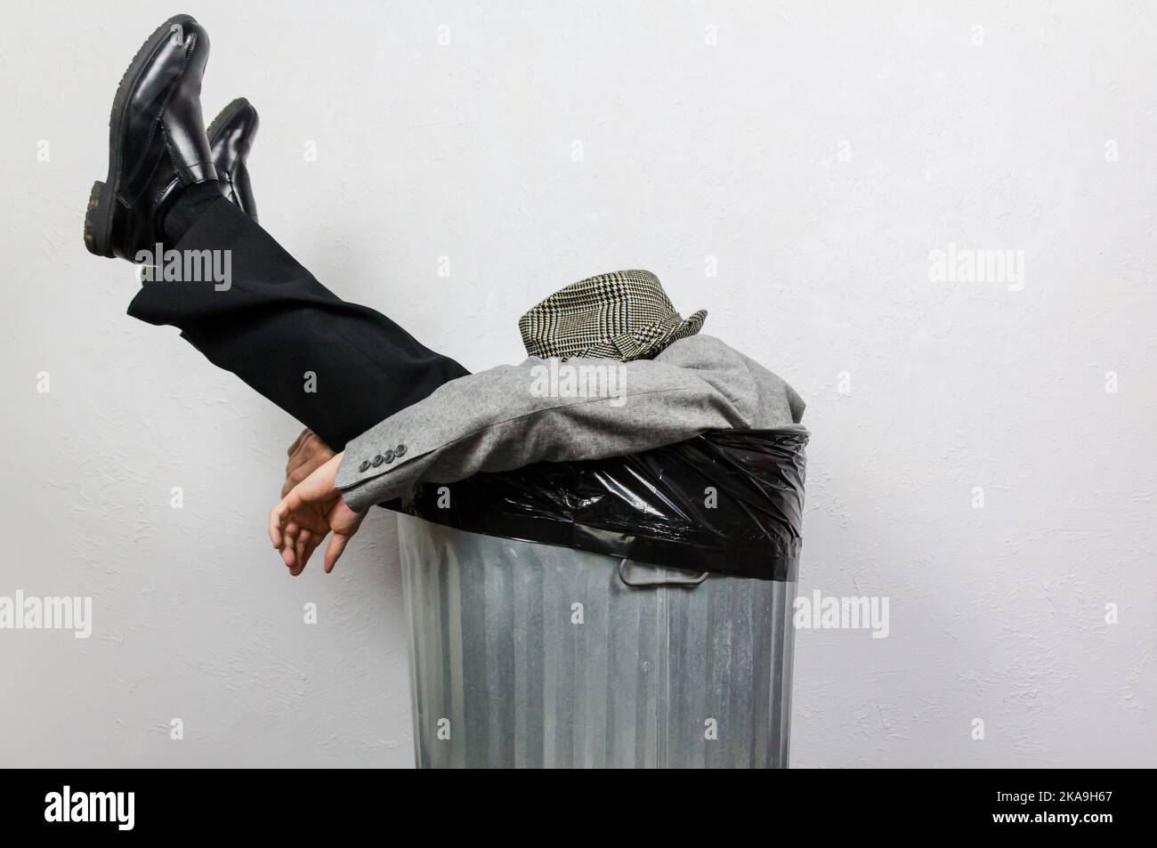 Portrait of Businessman in Suit and Fedora Hat Sitting in Trash Can. Man Thrown Away by Capitalism and Greed. Stock Photo