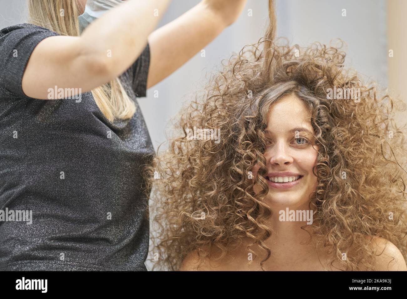 The hair master in face mask makes a curly hairstyle for a smiling woman. Portrait of a beautiful Caucasian girl with flowing curly hair. The concept of beauty salon. High quality photo Stock Photo