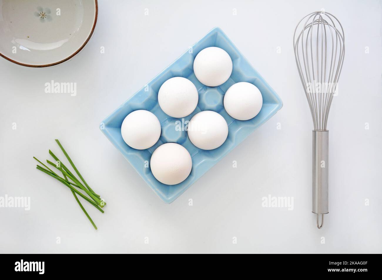 Farm fresh white jumbo eggs in blue ceramic egg carton with whisk, chives and bowl in flat lay composition.  Healthy ingredients on white background Stock Photo