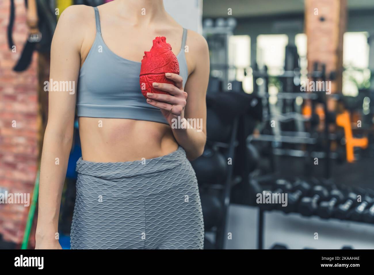 Conceptual gym photo. Pro-health physical activities. Unrecognizable caucasian person in gray gym clothes standing and holding vivid red fake heart in front of chest. High quality photo Stock Photo