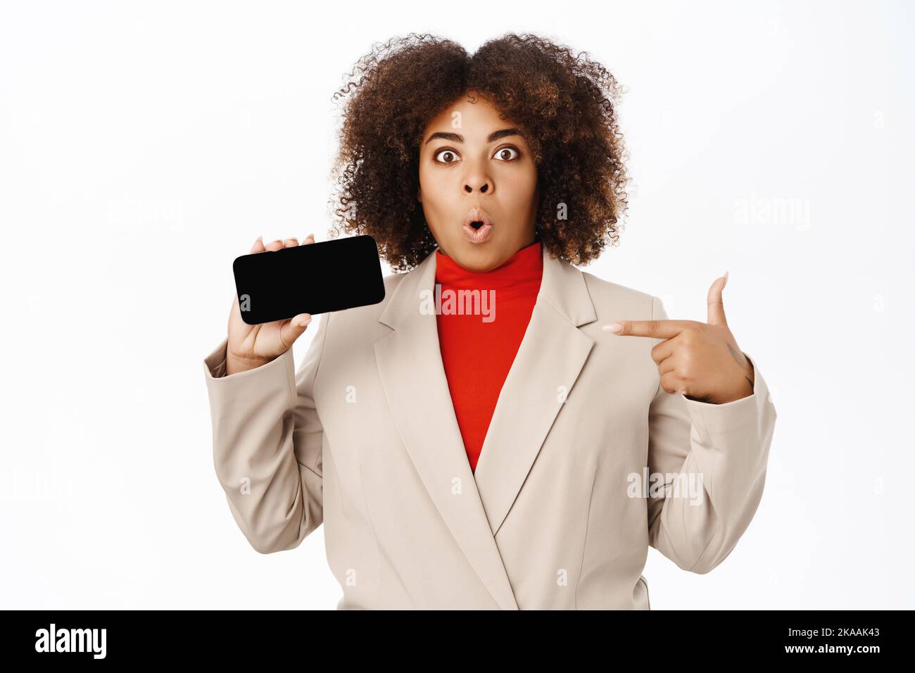 Portrait of businesswoman points at horizontal smartphone screen, shows app interface, company application or online store, stands over white Stock Photo