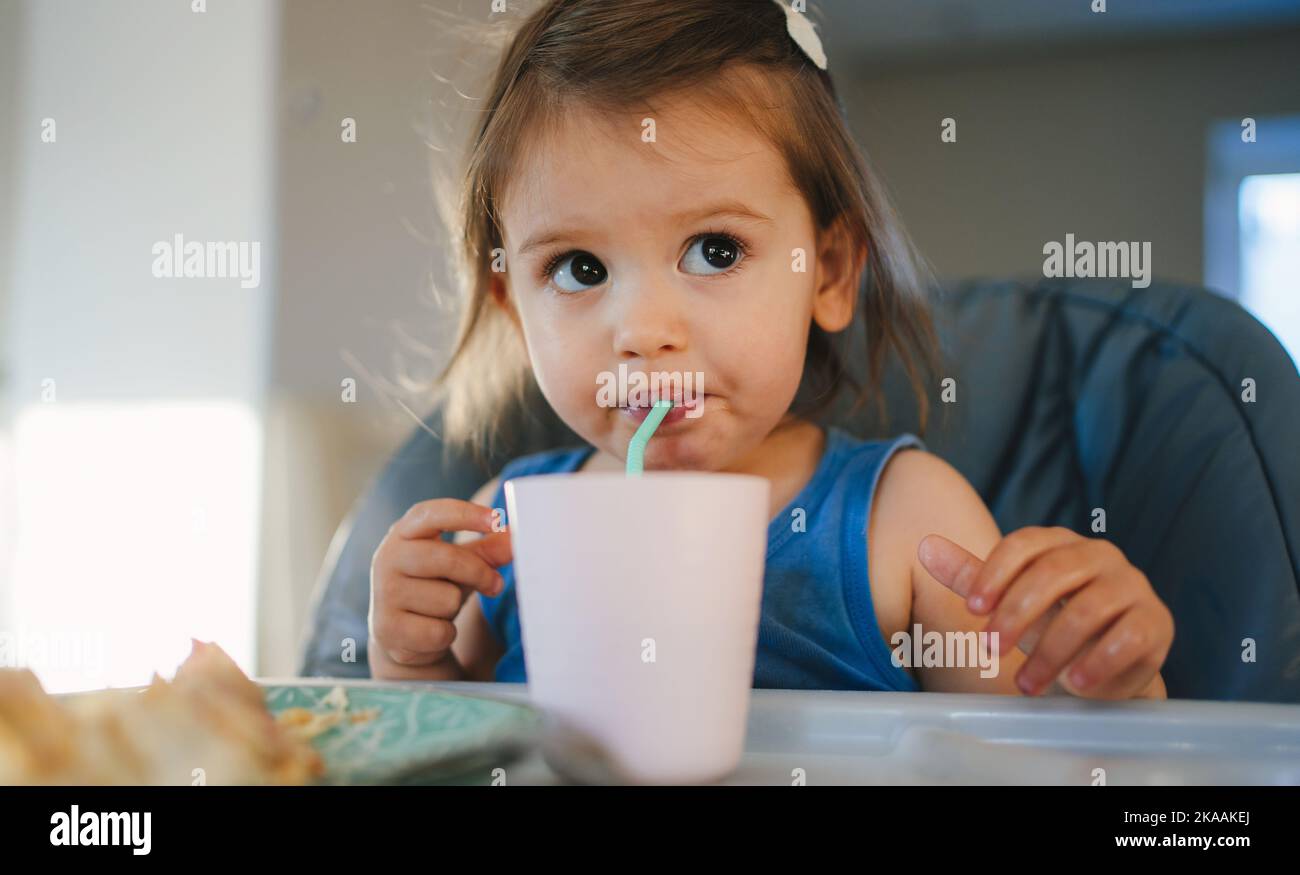 Baby girl drinking by herself with straw sitting in high chair at home. She is holding the