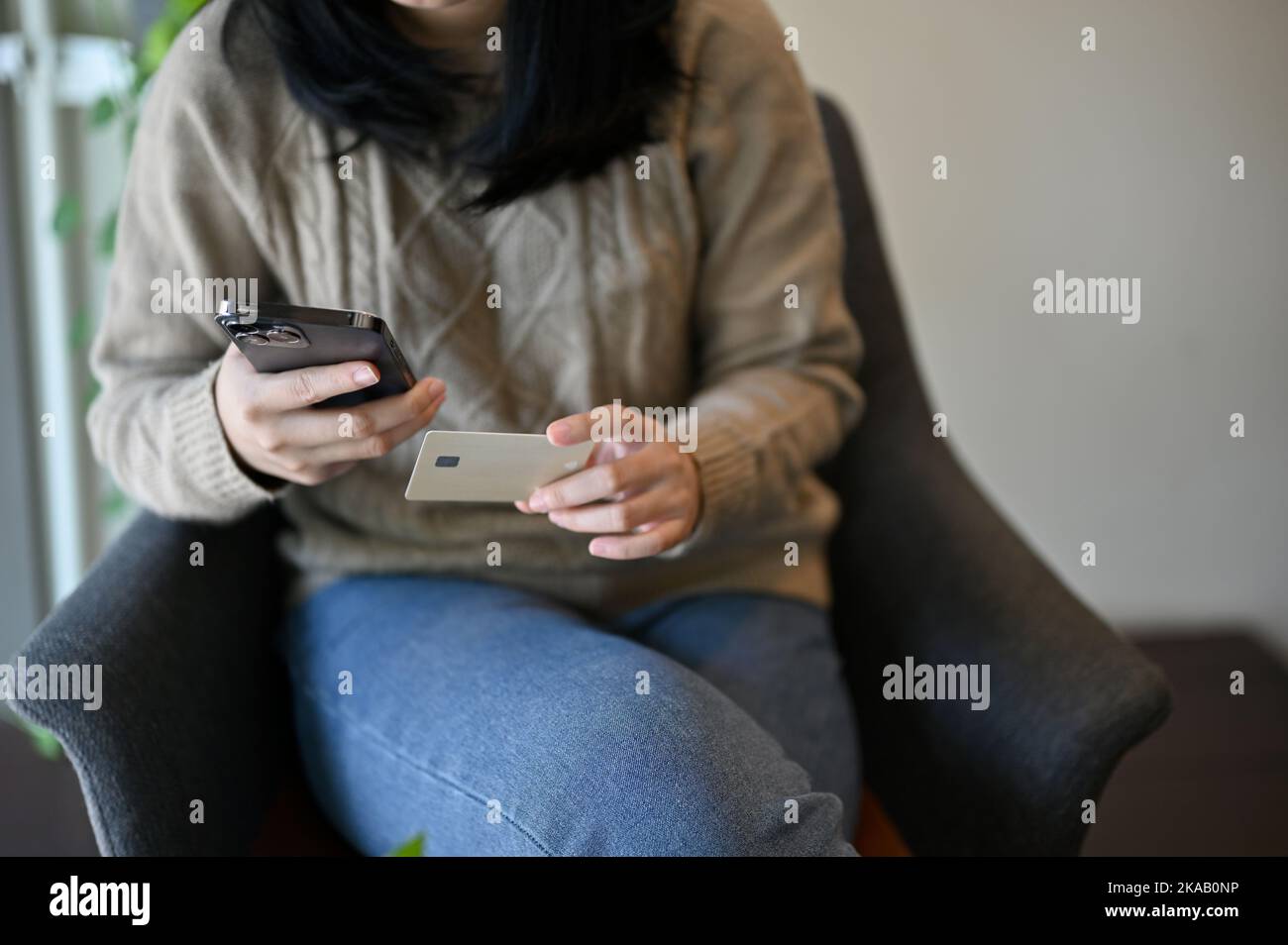 Attractive young Asian woman sitting on armchair, holding a credit card and her phone, paying