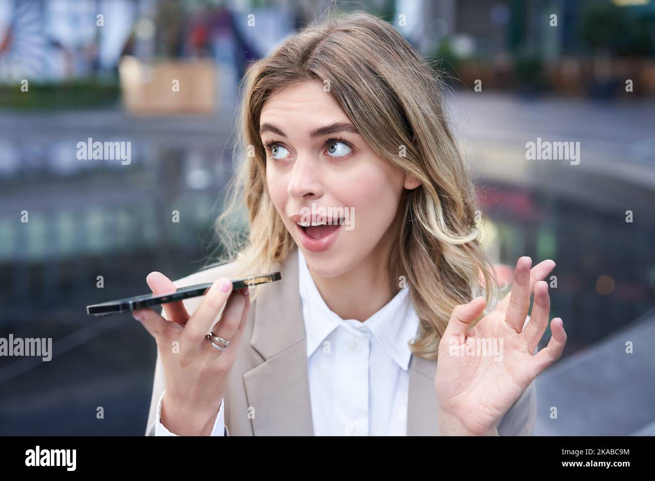 Chatty businesswoman sitting in city centre and recording voice message, talking into microphone on smartphone Stock Photo