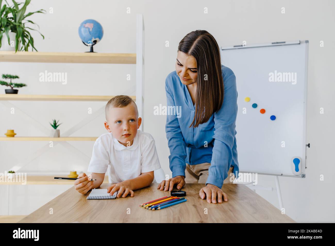 Private lesson. Attentive young woman tutor teacher helping little boy pupil with studying, correct mistakes explain learning material. Smiling mother Stock Photo