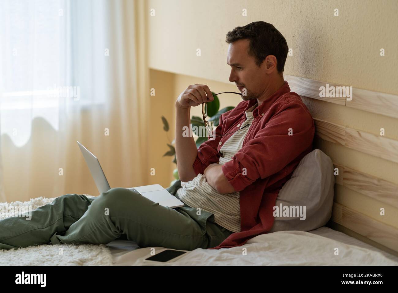 Young man freelancer sits on bed in hotel room with laptop on lap keeping track of new job orders  Stock Photo